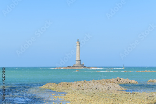 A tall stone lighthouse stands alone on a rocky outcrop surrounded by clear turquoise water and gentle waves beneath a cloudless blue sky near Cap de la Hague. The scene feels bright, open, and