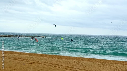 Wide static shot of kitesurfers and windsurfers riding winter waves off a sandy Barcelona beach under a cloudy Mediterranean sky.