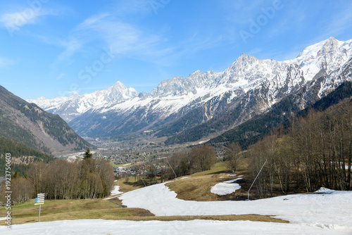 Sweeping view over the Chamonix valley with scattered spring snow patches, bare trees, and the dramatic snow-capped Mont Blanc massif under a bright blue sky.