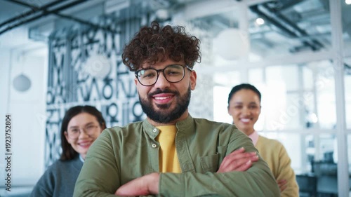 Modern office interior with three colleagues standing in bright workspace. Man in foreground smiling with arms crossed. Confident team celebrating successful collaboration and project completion.
