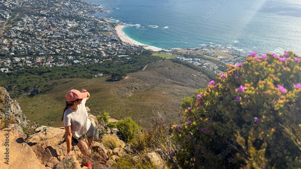 Fototapeta premium A hiker enjoys the panoramic view from Lion's Head in Cape Town, South Africa, overlooking the bay of Camps Bay and the surrounding coastline.