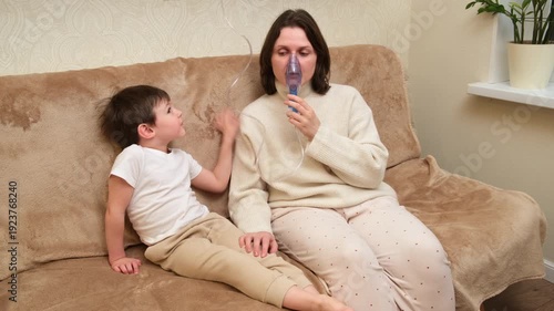 Woman making inhalation with nebulizer mask on couch next to young child. Home treatment respiratory disease for cold, cough, bronchitis, asthma.