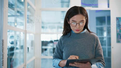 Charming female employee walking along office corridor and tapping on tablet screen. Woman wearing glasses and focusing on digital task. Modern corporate interior surrounding busy person.