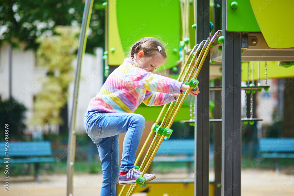Fototapeta premium Little girl climbing playground equipment using ropes and platforms in a green city park