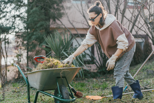 Obraz na plátne Young woman putting dry grass and leaves into wheelbarrow while cleaning backyard in spring