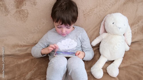 Child boy using tablet laying on sofa with stuffed toy. Kid enjoying digital content, screen time, gadget, early childhood education concept.