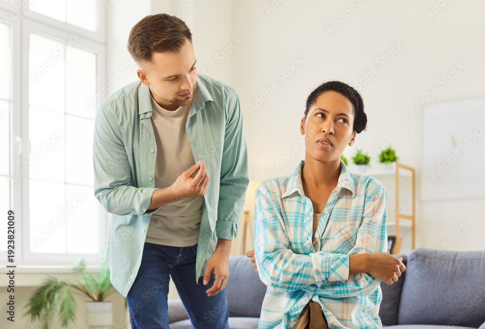 Fototapeta premium Couple conflict at home, man speaking and gesturing towards woman wife sitting on sofa, turning away head, crossing arms in annoyance, ignoring, domestic abuse, disagreement, frustration and anger
