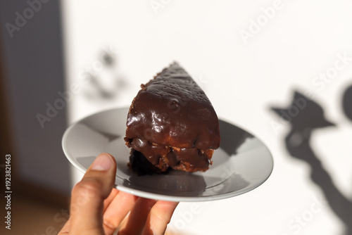 Hand holding a plate with a piece of dark chocolate cake. Conceptual food photography with deep shadows on a white background.