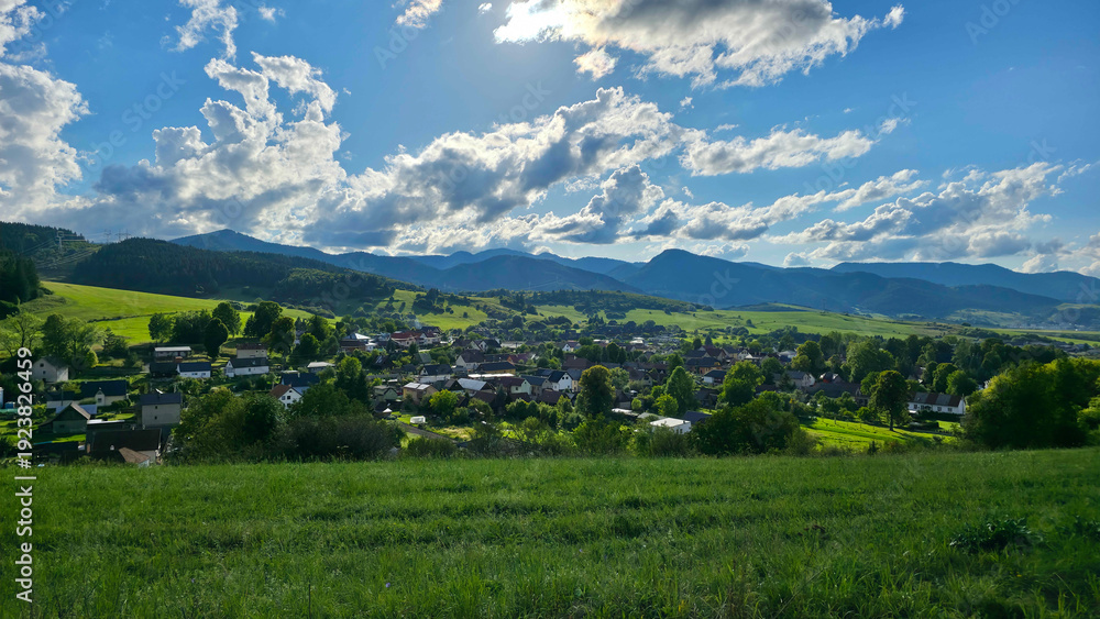 Fototapeta premium Picturesque landscape view of a peaceful rural village nestled in a vibrant green valley, surrounded by rolling forested mountains under a cloudy sky.