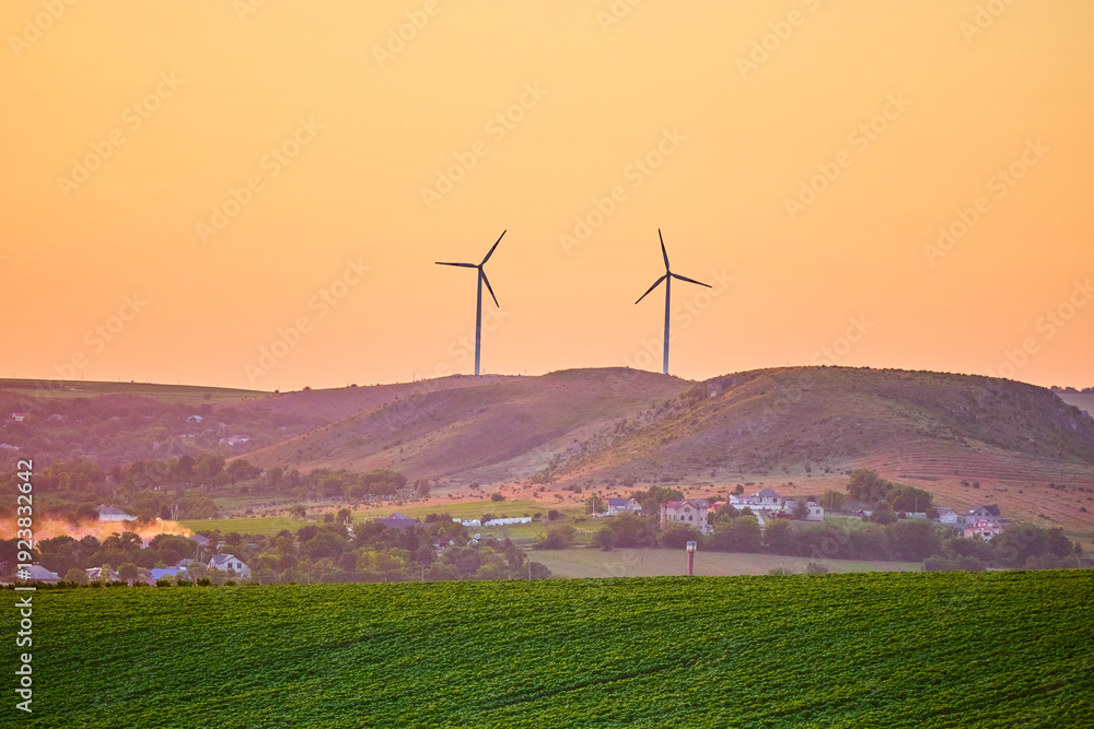 Obraz premium Wind turbines on rolling hills at sunset with farmland and orange sky