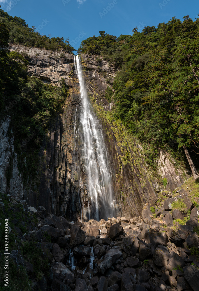 Obraz premium A dramatic waterfall pours down a sheer stone face surrounded by lush green forest at Nachi Falls in Wakayama, Japan. Sunlight enhances the rugged textures of the rocks and the vibrant foliage under a