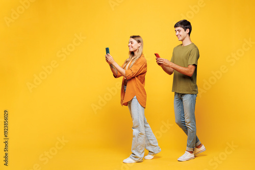 Photography Full body sideways fun young couple two friends family man woman wear casual clothes hold in hand use mobile cell phone together isolated on pastel plain light yellow color background studio portrait