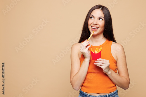 Young minded woman wear orange casual clothes hold box with french fries potato look aside area isolated on plain pastel beige background. Proper nutrition healthy fast food unhealthy choice concept.