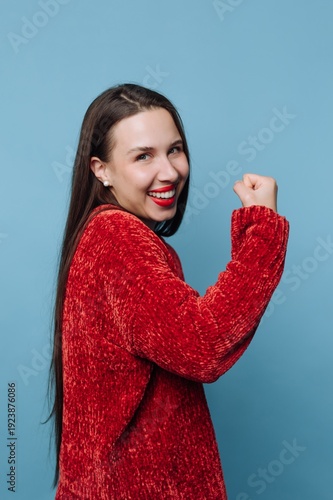 Slika na platnu Joyful woman in a red sweater flexes her arm with a confident smile against a vi