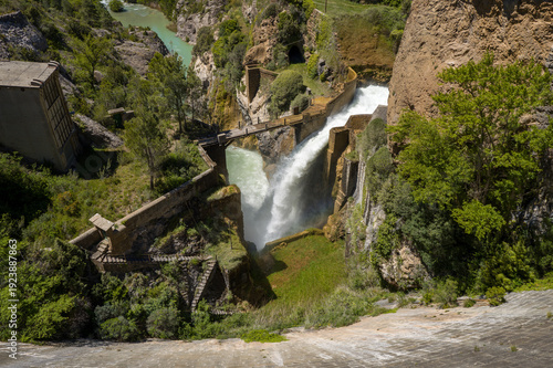 Canvas Print A dramatic waterfall cascades from a dam outlet into a rocky gorge at Las Penas, surrounded by lush greenery, rugged cliffs, and turquoise river water