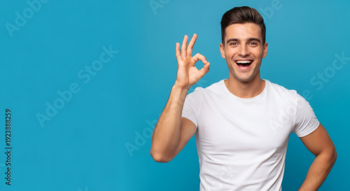 Young man in white t-shirt showing OK gesture and smiling broadly against blue background
