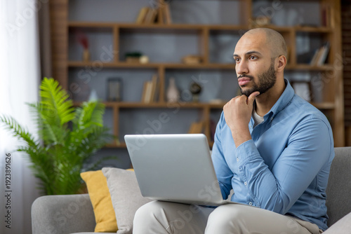 Fotografi Young adult man on a sofa with a laptop, looking away while contemplating ideas,