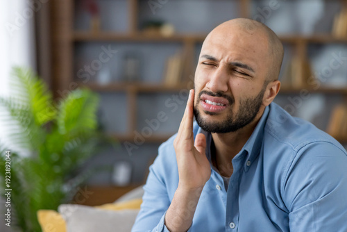 Young man in a blue shirt clutching his cheek in a living room, grimacing from severe tooth pain and seeking urgent dental care with a pained, distressed expression