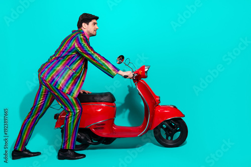 Energetic man in a rainbow suit rides a red scooter against a bright turquoise backdrop for a bold lively photo shot