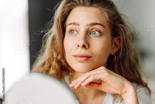 Принти на полотні A close-up of a young woman touching her face and looking into a small mirror on a sunny day
