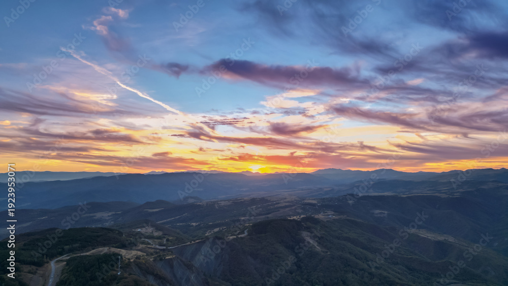 Fototapeta premium Aerial panoramic view of Georgia at sunset with golden-orange sun, colorful layered clouds, vast mountain ranges, forested slopes, and winding mountain road