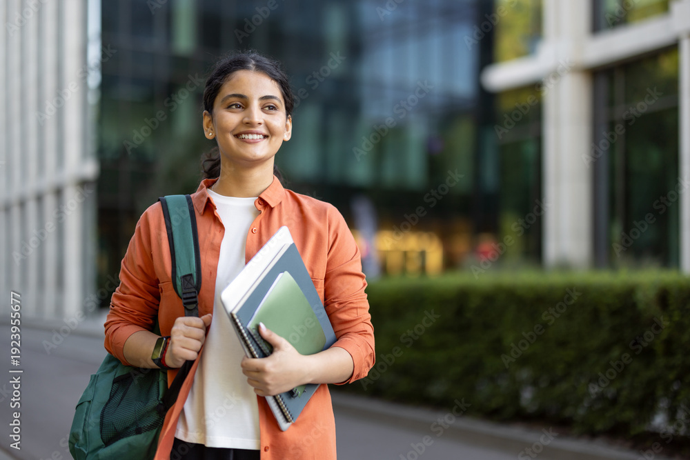 Fototapeta premium Happy indian student carrying a backpack and notebooks, confidently walking on a university campus, representing education, success, and future career aspirations