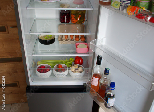 Open refrigerator interior, fresh vegetables, fruit, eggs, ham, cheese and homemade pickles on clean glass shelves. Oganized fridge drawer with mushrooms, radishes, tomatoes, celery and carrots, eggs.