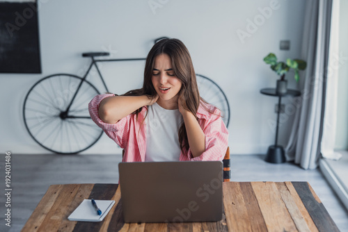 Картината върху платно A tired woman sits at a table rubbing her neck and tense shoulder muscles, suffe