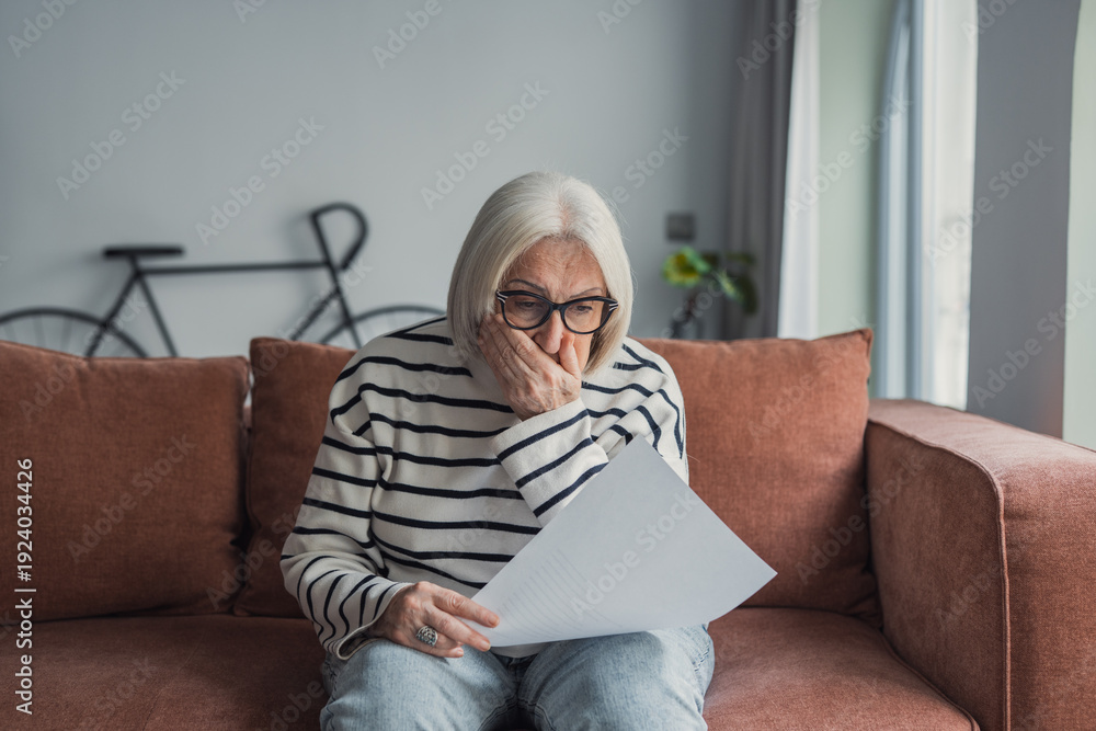 Fototapeta premium Upset elderly woman sitting on couch at home, reading document and getting stressful bad news at home. Frustrated senior lady shocked with letter, touching head, trying to cope with negative emotions