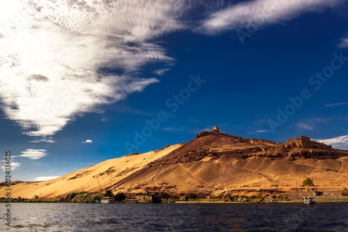 Fotografija Golden sand dunes slope down to a calm Nile river where small boats moor beneath ancient ruins on the hillside
