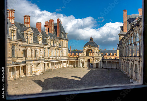 Window-framed interior view onto the courtyard of Château de Fontainebleau in France, showing historic façades, arches, and a central pavilion beneath dramatic clouds and blue sky, UNESCO Site