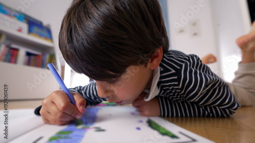 Young boy lying on the floor, drawing with a blue marker, deeply immersed in creative activity, focused expression, engaged in artistic learning and imagination at home