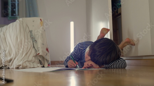Young boy lying on the floor, resting his head on his arm while holding a green marker, appearing tired or lost in thought, taking a break from drawing in a cozy home setting