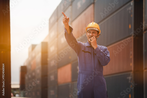 Logistics worker using walkie talkie and pointing in container yard, coordinating cargo and shipping at industrial port, concept of teamwork, safety, transportation and global supply chain.
