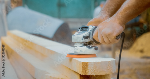 Hands are working skillfully to sand wooden beams with an angle grinder, creating smooth surfaces in a busy construction area. Dust clouds fill the air as the job progresses.