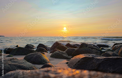 A serene sunset seascape. Granite rocks on a sandy shore catch the golden light as the sun descends toward a calm sea