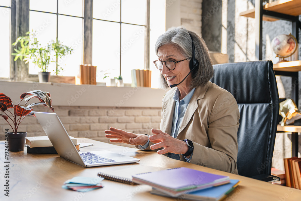 Fototapeta premium Virtual communication. Smiling mature woman remote working interpreter tech support consultant in headset with mic talk to client via video call conference app on pc engaged in online event.