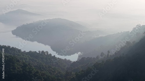 Mountain range with a lake in the foreground and a foggy sky in the background. The mountains are covered in trees and the lake is calm