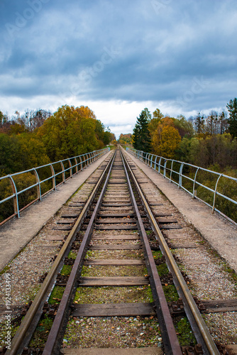 Railway tracks of two different gauges run parallel on an old narrow bridge surrounded by autumn foliage. The sky is overcast, adding a dramatic effect to the landscape.