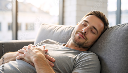 Young man sleeping on a grey sofa in a bright living room. Relaxed male taking a nap at home. Wellness and rest concept