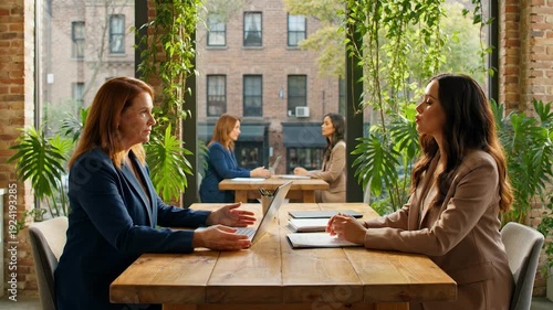 Two businesswomen in a modern office setting with plants and laptops on a wooden desk