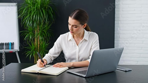 Businesswoman working on laptop at office desk with notebook and smartphone nearby