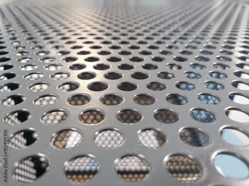This high-angle, close-up photograph features a grey perforated metal plate. The surface is defined by a repetitive, geometric grid of uniform circular holes. 