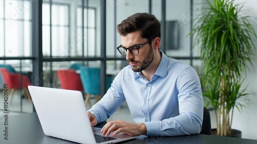 Man sitting at a desk working on a laptop in a modern office environment with a plant