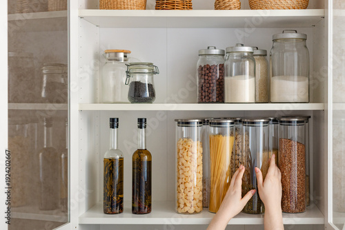 A woman takes a jar from a kitchen shelf filled with food containers and glassware. The cabinet door is open.