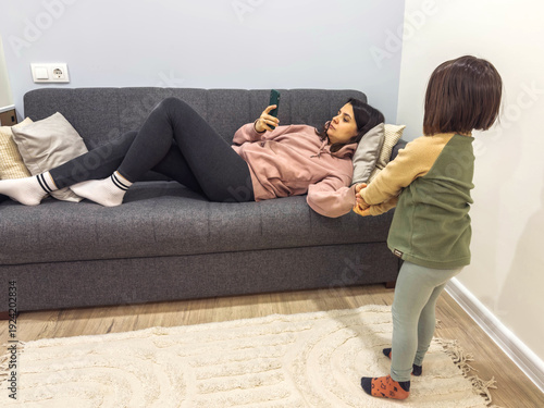 Photography Mother lying on sofa using smartphone while little child stands nearby holding h