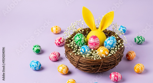 Festive Easter still life: decorative eggs, small white flowers, bunny ears in a woven nest on light purple surface, embodying spring holiday charm