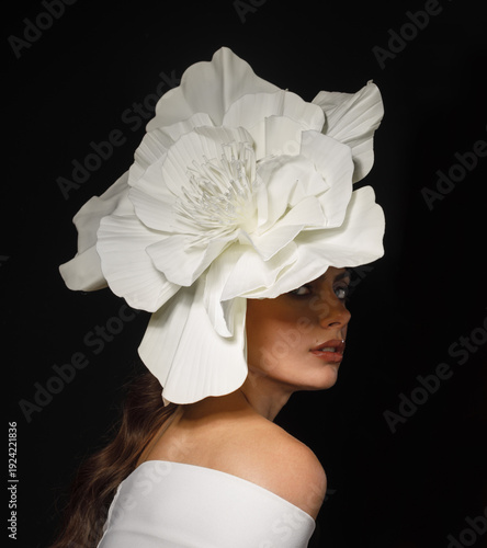 Portrait of woman wearing oversized white flower headpiece on black background. High fashion, creative styling and dramatic beauty concept.