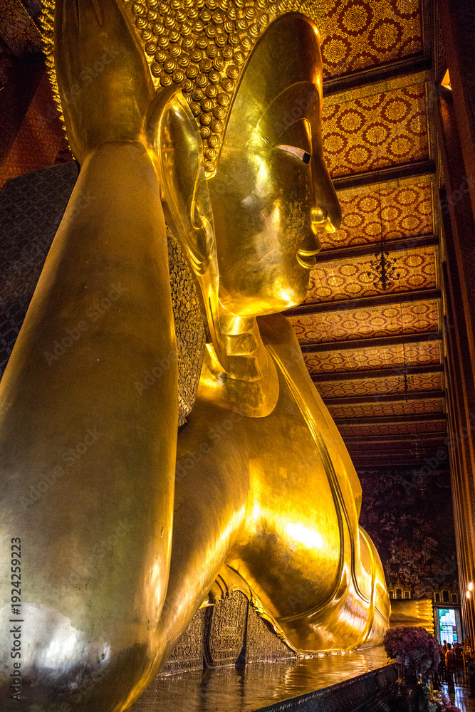 Obraz premium Close up of the colossal Reclining Buddha statue at Wat Pho in Bangkok, Thailand. The entire statue is covered in brilliant, reflective gold leaf that glows under the temple's warm lighting.