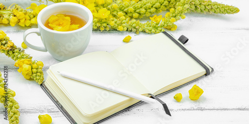 Cup of herbal tea with mullein flowers and blank notebook with pen on white wooden table